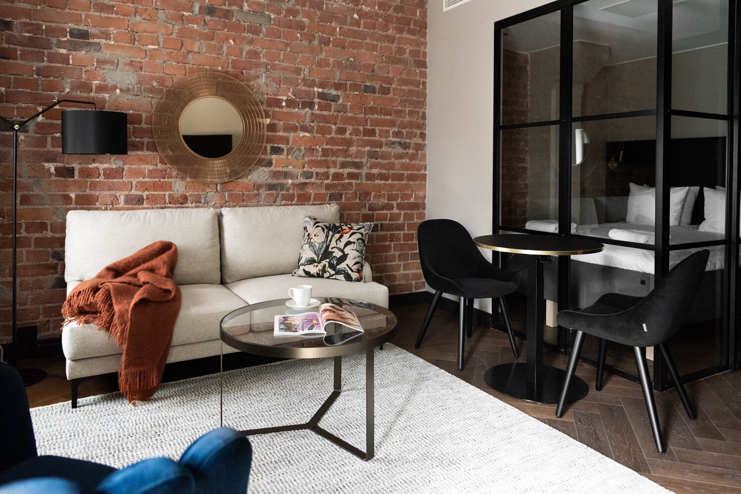 A modern living room with a brick wall, a round mirror, a sofa, a coffee table, and a glass partition separating the living area from a bedroom.