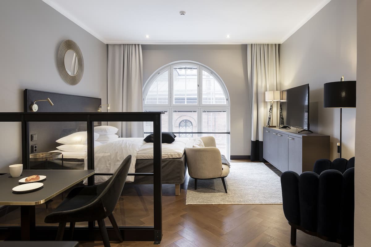 A modern hotel room with a bed, a desk with a chair, a TV, and a large window.