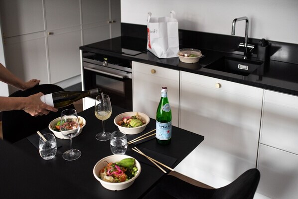 Una cocina moderna con encimera negra, armarios blancos y un lavamanos de acero inoxidable. Se observan cuencos con comida, vasos y una botella de agua S.Pellegrino.