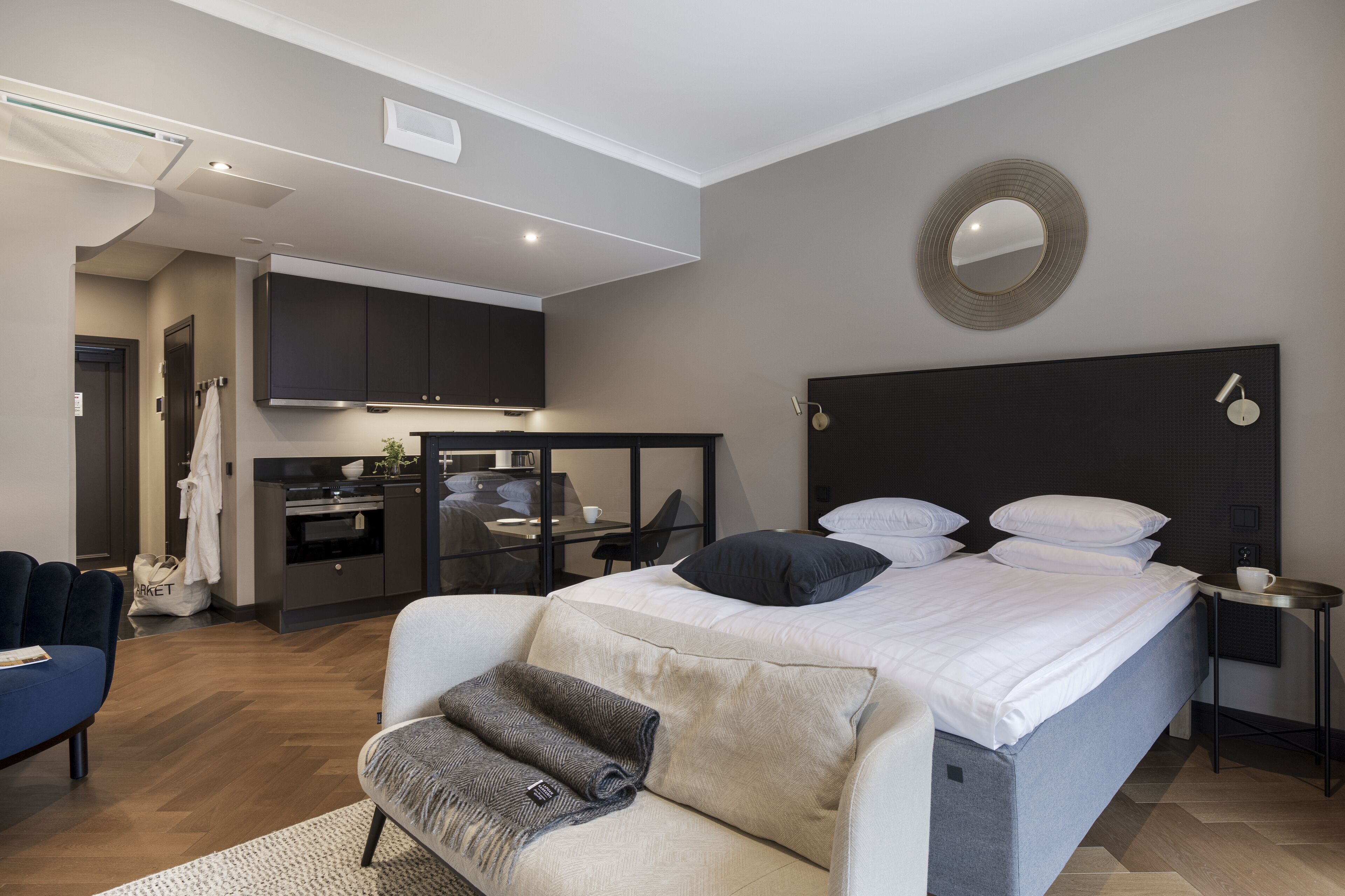 A modern hotel room with a bed, a sofa, a small table, a mirror, and a kitchenette.