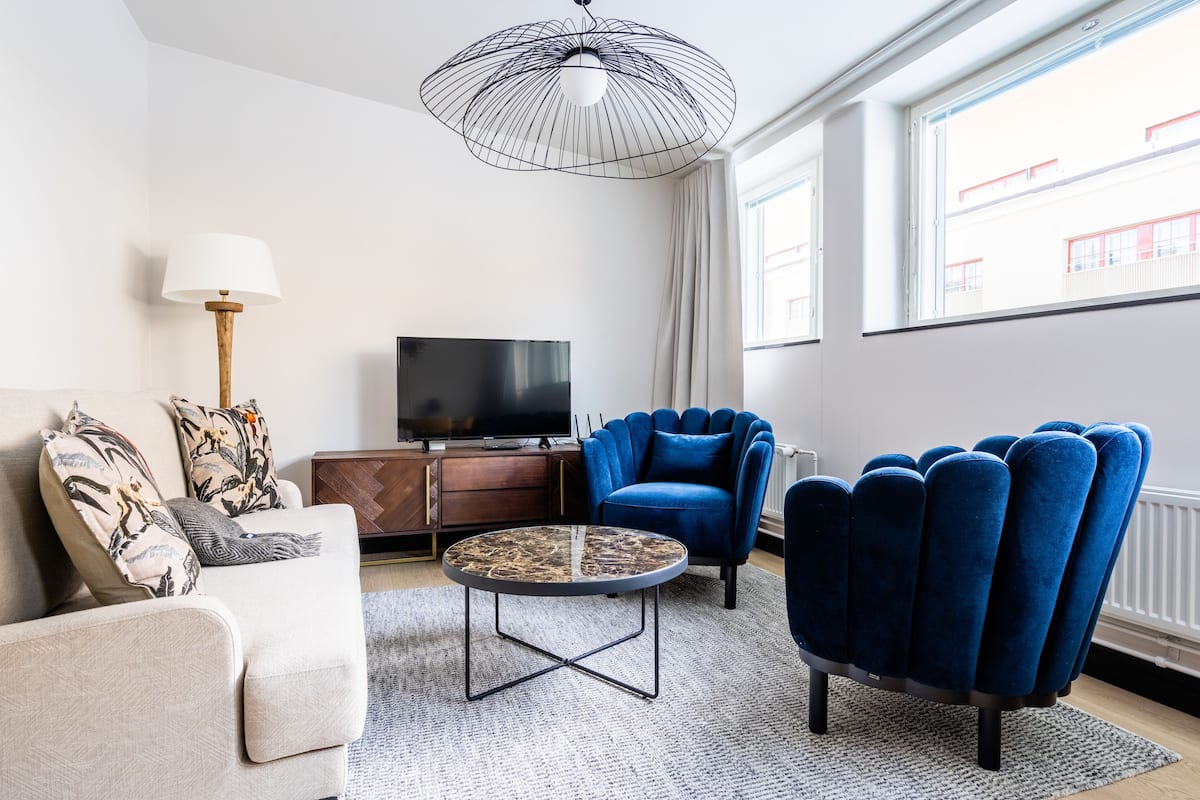 A modern living room with a sofa, a blue armchair, a coffee table, a TV, and a large window with a view of buildings.