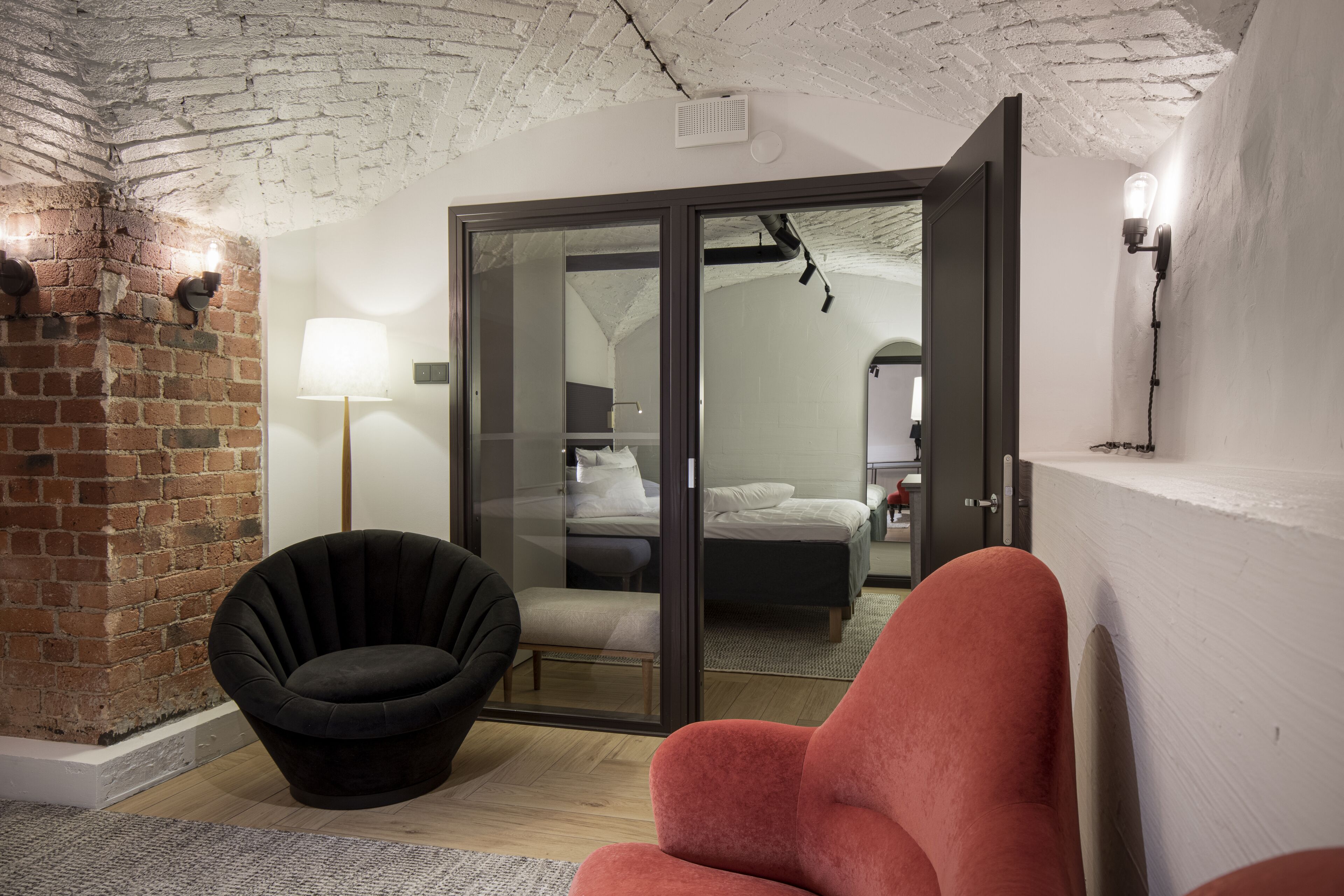 Una habitación moderna con pared de ladrillo, una silla negra, una silla roja, un ventanal de vidrio que da a un dormitorio y una lámpara en la pared.
