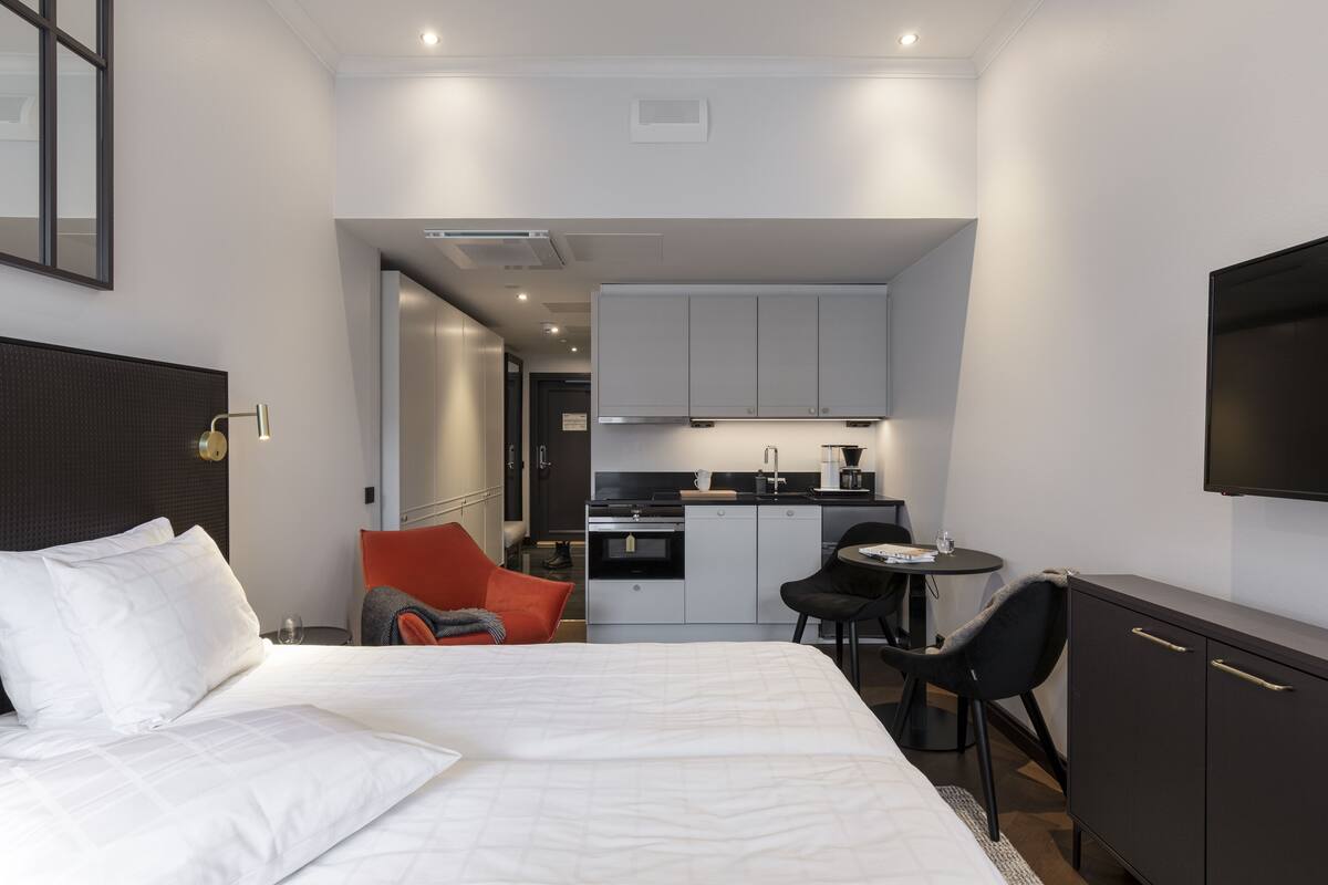 A modern hotel room with a large bed, a desk, a chair, a small kitchenette, and a TV mounted on the wall.