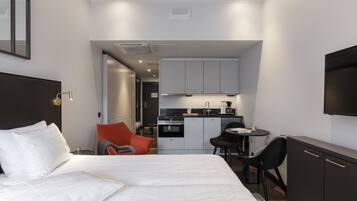 Quarto de hotel moderno com uma cama grande, uma escrivaninha, uma cadeira, um pequeno espaço com cozinha e uma televisão na parede.
