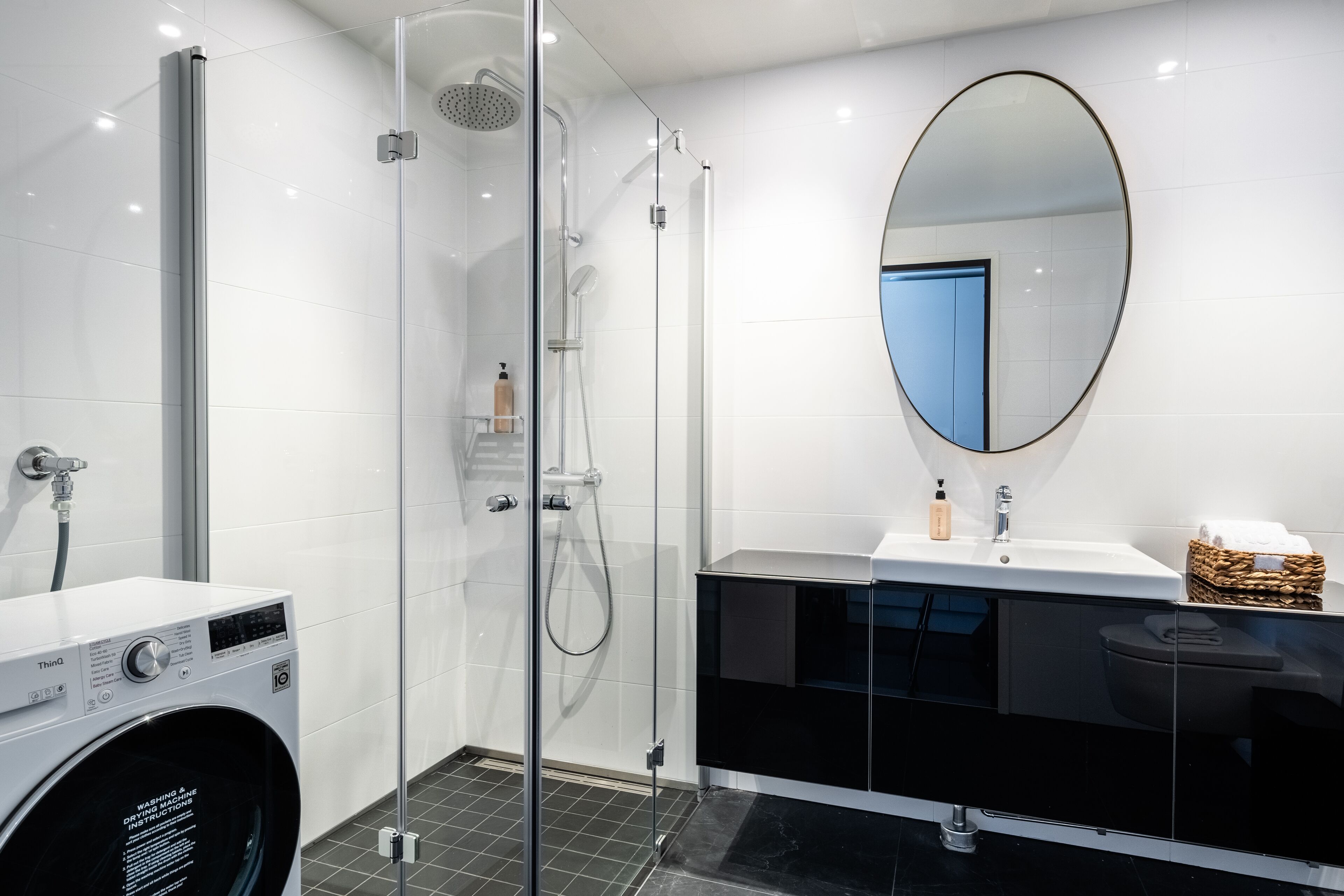 Un baño moderno con ducha de vidrio, lavadora blanca, tocador negro con espejo y piso de baldosas.