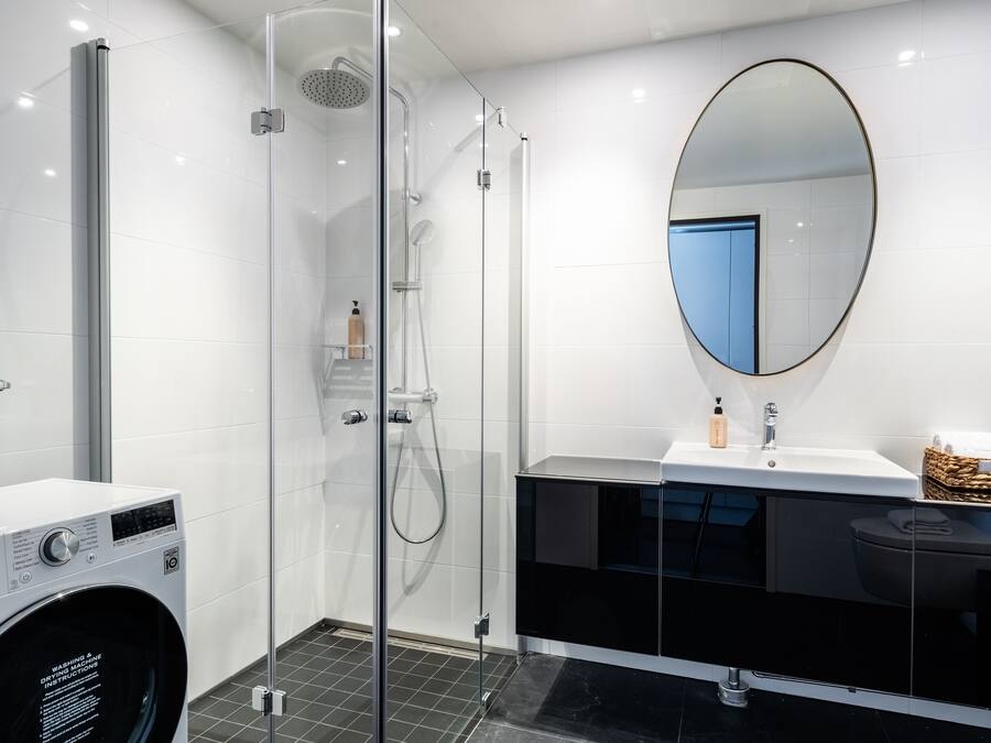 Un baño moderno con ducha de vidrio, lavadora blanca, tocador negro con espejo y piso de baldosas.