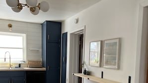 A modern kitchen with wooden flooring, dark blue cabinets, a built-in dishwasher, and a ceiling fan. There are two wooden stools and a window with a view of greenery. - Historic Hudson Valley 1850 Brick Home (Saugerties)