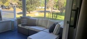 A covered porch with a beige sectional sofa, a small table, and a view of a grassy yard and trees. - Villa Tortuga Modern Updated home with 2beds/2bath  Modern Farmhouse w/Golfcart
 (The Villages)
