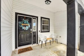 A front porch with a black door, a welcome wreath, two white chairs, and a doormat.