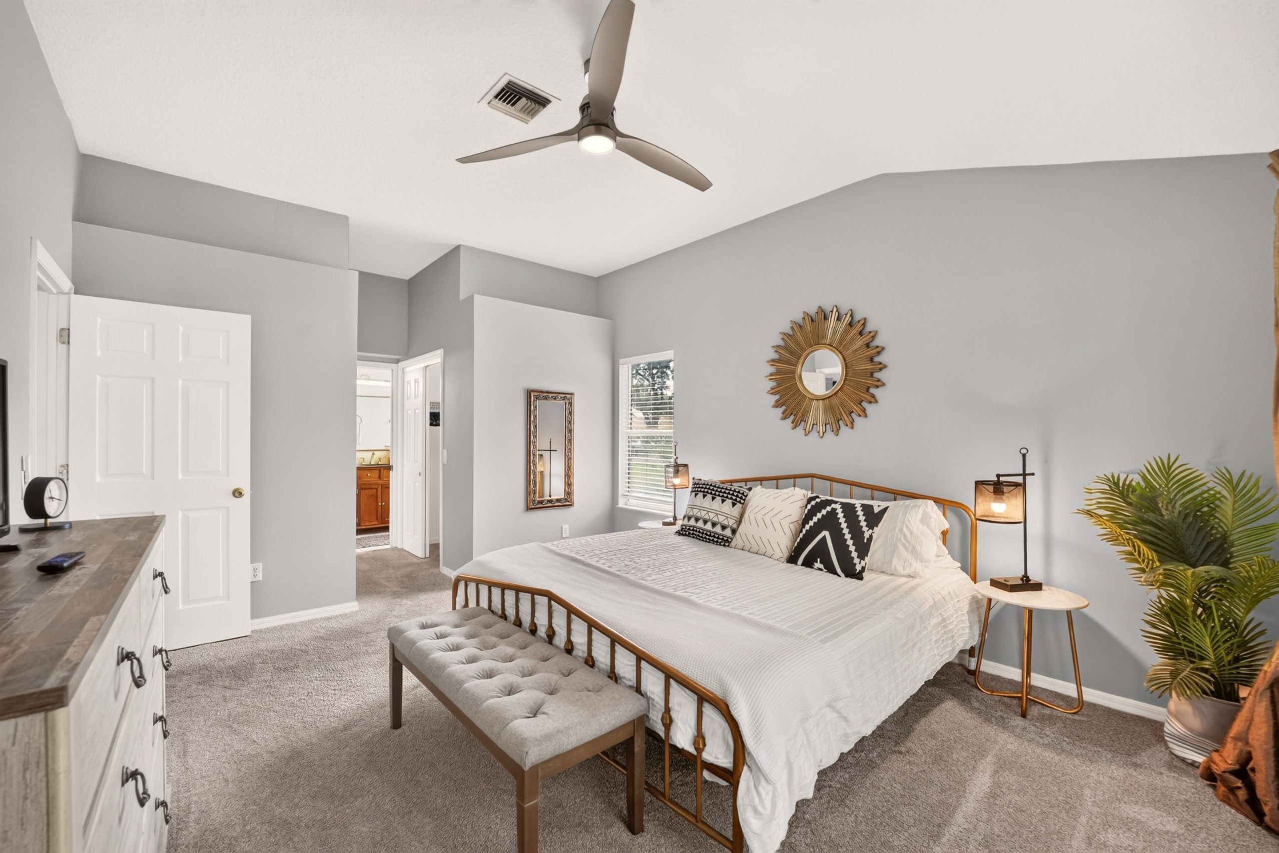 A bedroom with a bed, a bench, a ceiling fan, a wall-mounted mirror, a bedside lamp, and a potted plant.