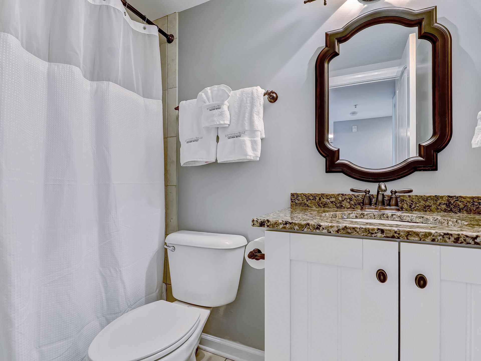 Une salle de bain comprenant des toilettes, un lavabo avec un plan de travail en granit, un miroir avec un cadre en bois et un rideau de douche.