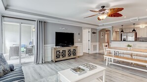 A modern living room with a flat-screen TV, a ceiling fan, and a dining area with a wooden table and chairs. - Spacious Condo, Walk to Beach, Heated Pool Access (Tybee Island)