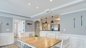 A dining area with a wooden table and white chairs, a kitchen with a refrigerator and stove, and a hallway with a door leading to another room. - Spacious Condo, Walk to Beach, Heated Pool Access (Tybee Island)