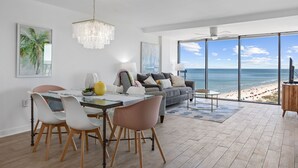 Ett modernt vardagsrum med ett stort fönster med utsikt över en strand, en matplats med ett glasbord och en mysig sittgrupp.