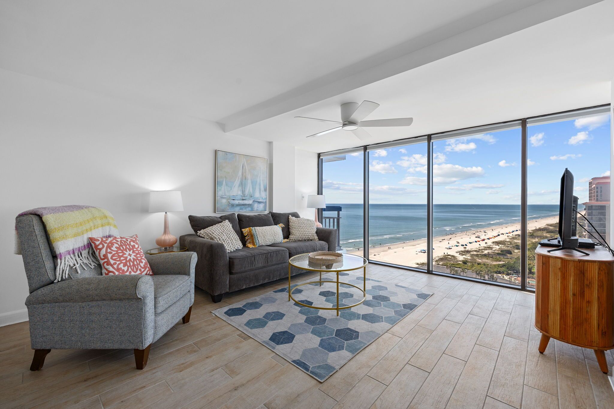 Ein modernes Wohnzimmer mit Blick auf den Strand, ein grauer Sessel, ein graues Sofa, ein runder Couchtisch und ein Holznachttisch.