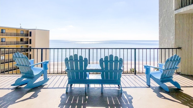 A balcony with two rocking chairs and a small table, overlooking a beach and a building with multiple balconies. - Top Of The Gulf 213 (Panama City Beach)
