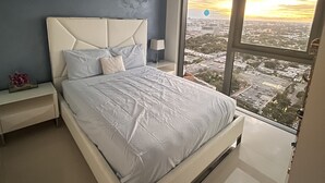 Un dormitorio moderno con una cama grande, una mesita de noche, una lámpara y vistas a un paisaje urbano a través de un ventanal.