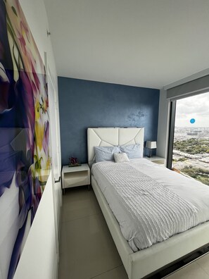 Un dormitorio moderno con una cama grande, una mesita de noche, una lámpara de pared y vistas a la ciudad a través de un ventanal amplio.