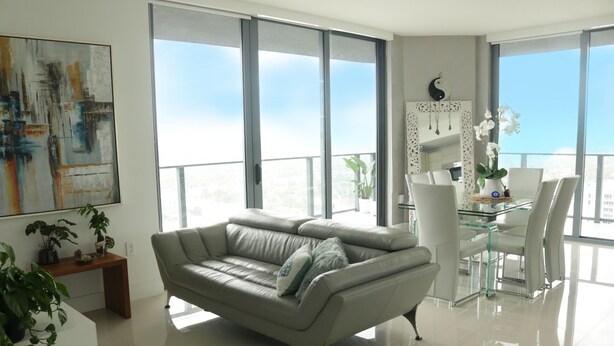 Un salón moderno con un sofá gris, una mesa de comedor de cristal y un ventanal con vistas a la ciudad.