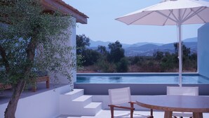 Une terrasse avec une table et des chaises, un parasol blanc, et une vue sur une piscine et des montagnes.