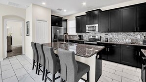 A modern kitchen with dark cabinets, a granite countertop, and a central island with bar stools. - Stunning 5 Bed Pool Home with Game Room-1585TL (Davenport)