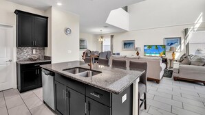 A modern kitchen with granite countertops, black cabinets, and a dishwasher. There is a dining area with a gray sofa and a TV. - Stunning 5 Bed Pool Home with Game Room-1585TL (Davenport)