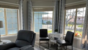 A room with a grey leather sofa, two black chairs, a small white table with a potted plant, and large windows with patterned curtains. - Charming home on the Cliffs of Crescent City (Crescent City)