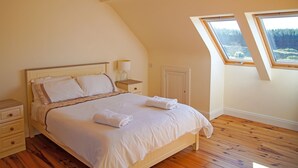 Une chambre avec un plafond en pente, un lit recouvert de draps blancs, une table de chevet avec une lampe et une fenêtre donnant sur des arbres.