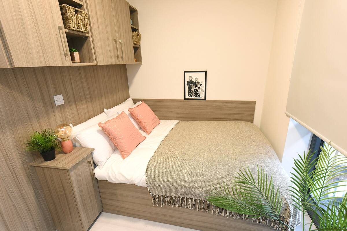 Ein ordentlich eingerichtetes Schlafzimmer mit einem Bett, Kissen, einem Nachttisch mit Lampe und einem Wandregal mit Körben.