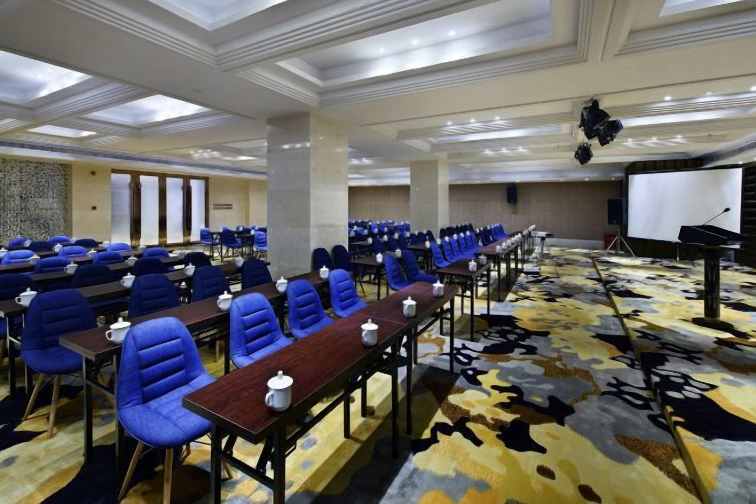 Un salón de conferencias con hileras de sillas azules, una larga mesa con micrófonos y un escenario con un atril.