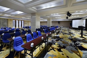 A conference room with rows of blue chairs, a long table with microphones, and a stage with a podium.