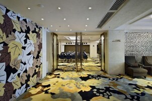Un vestíbulo de hotel con alfombra estampada, una sala de conferencias con mesas y una zona de estar con sillas.