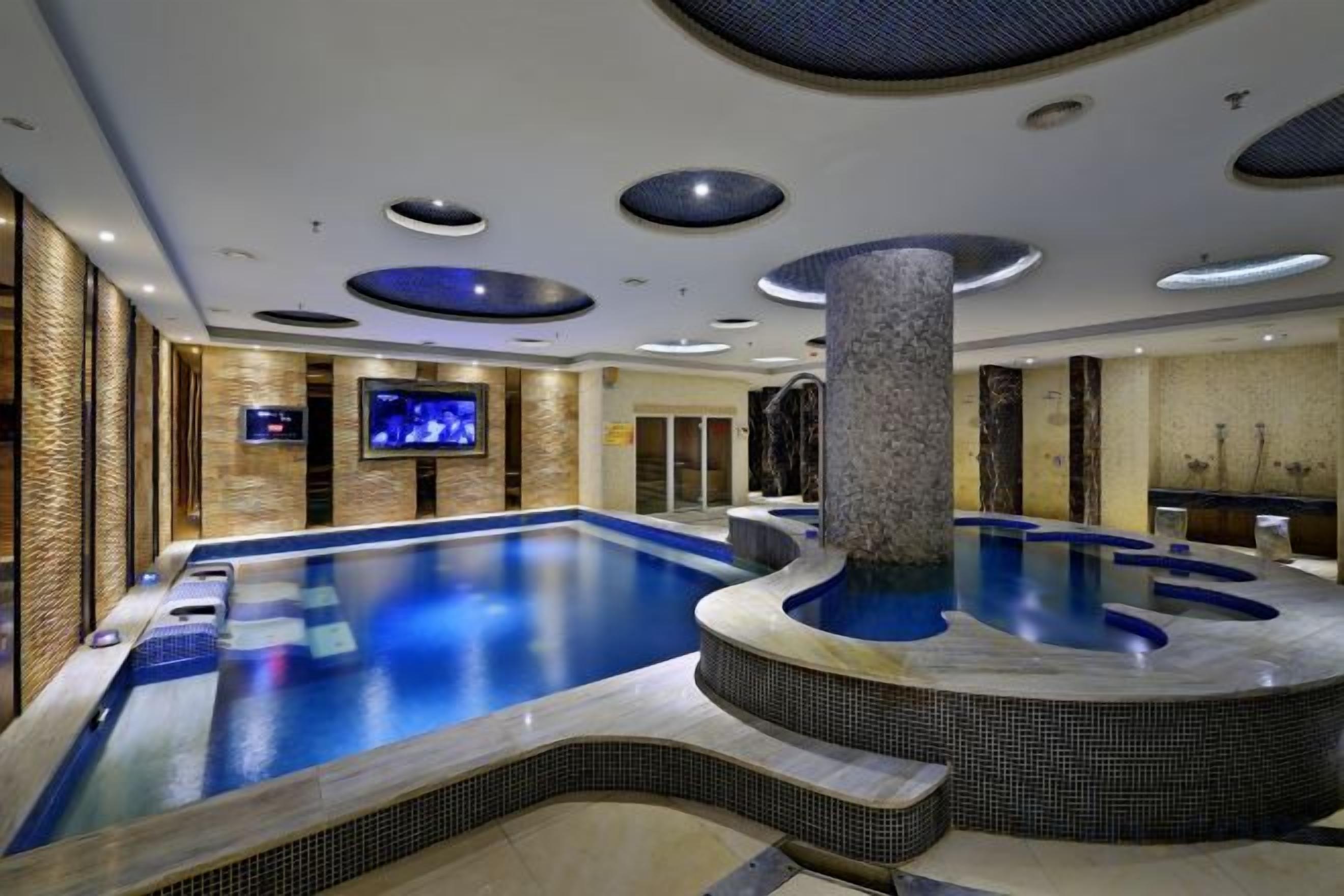 A large indoor swimming pool with a hot tub, a television screen, and a stone pillar.