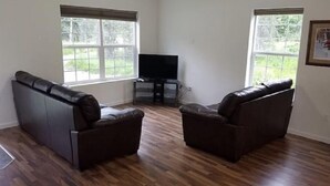 A living room with two leather sofas, a flat-screen TV on a stand, and large windows with a view of greenery.
