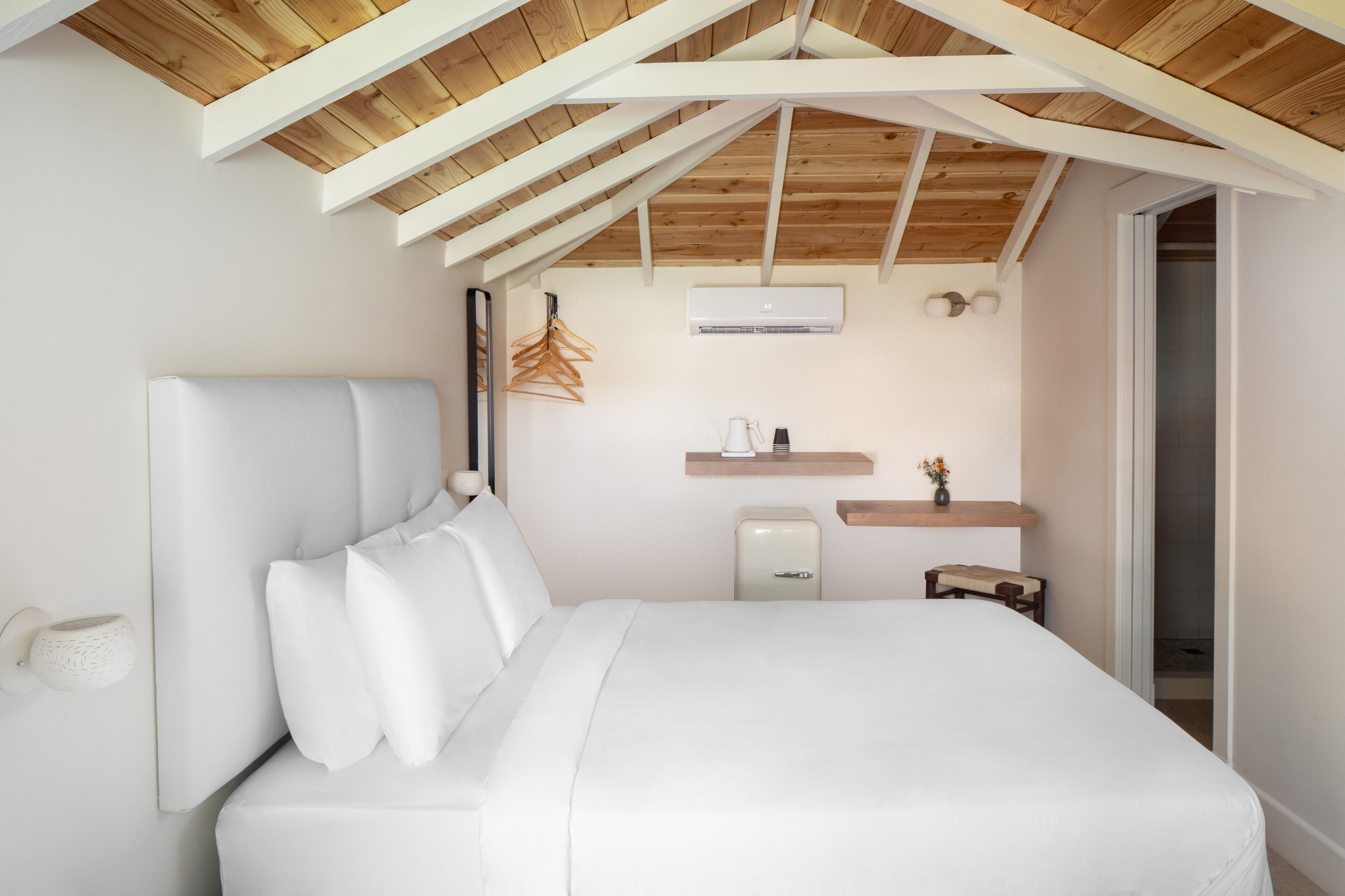 Un dormitorio con techo alto de madera, cama con ropa blanca, mesita de noche, pequeña lámpara de noche, aire acondicionado instalado en la pared y estante de madera con un jarrón.