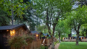 A row of wooden cabins with a deck and a small staircase, surrounded by trees and a well-maintained lawn.