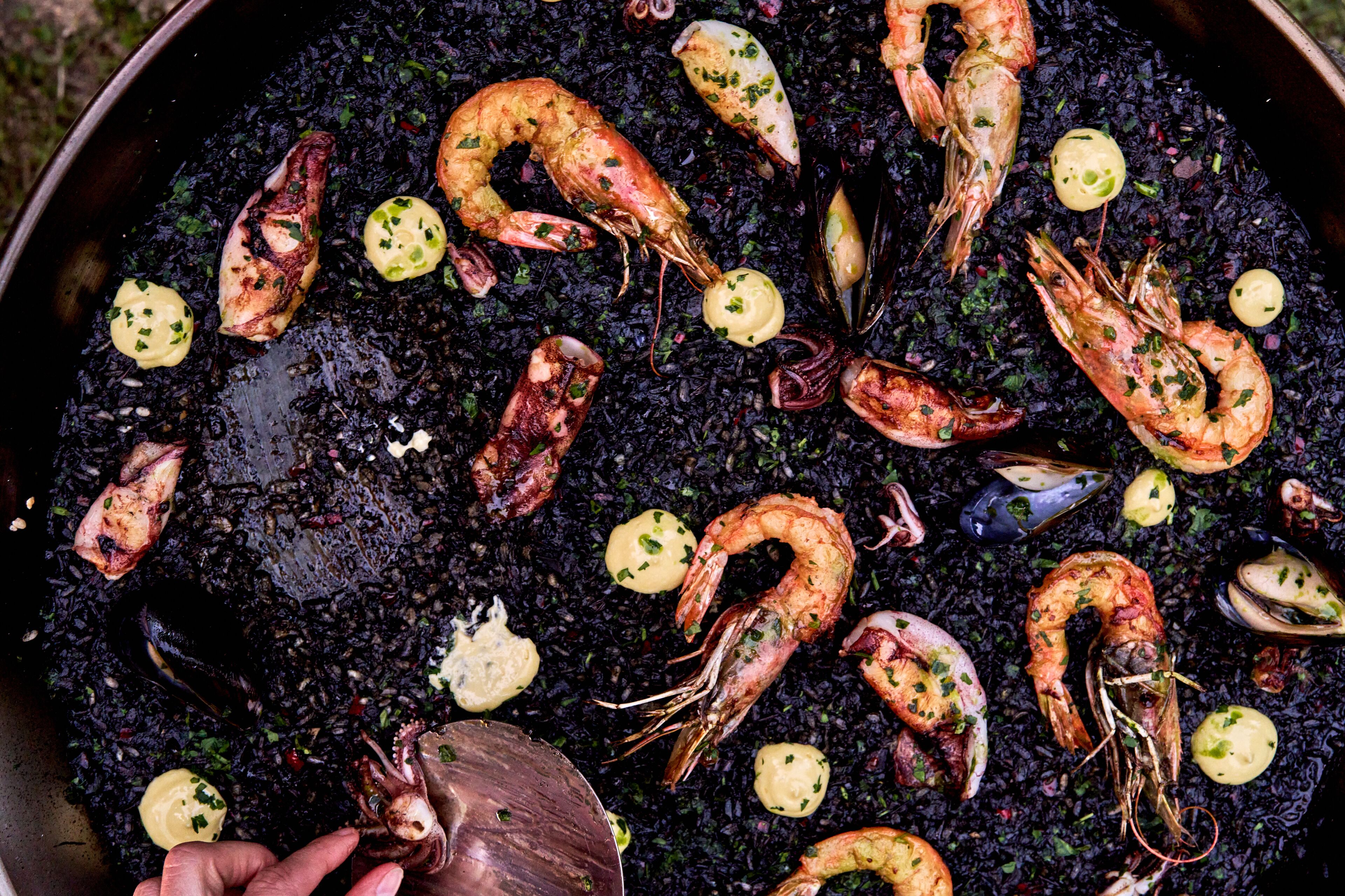 Una paellera grande llena de arroz negro, camarones y mejillones, decorada con hierbas y rodajas de limón.