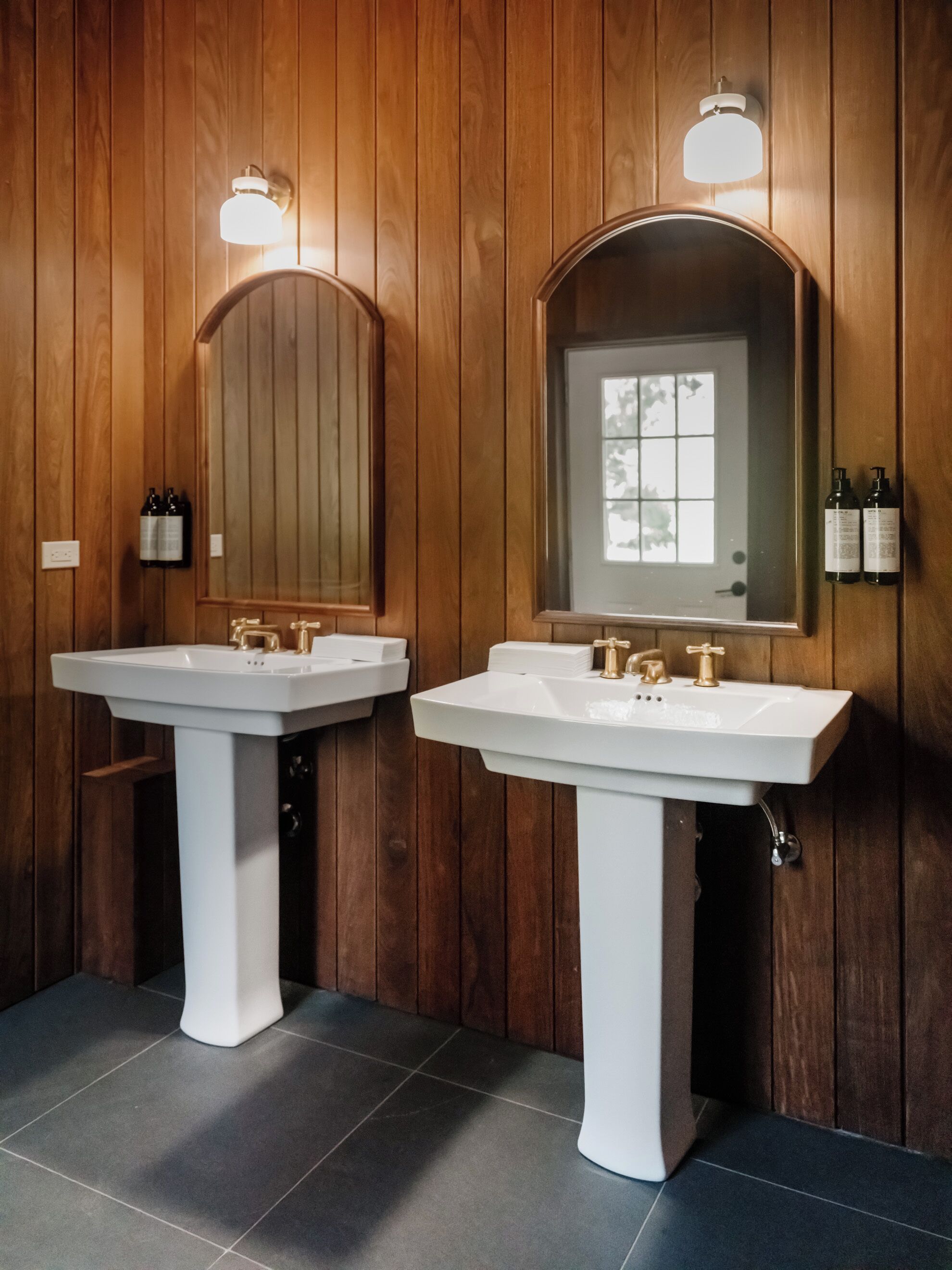 Deux éviers blancs, dotés de robinets dorés, sont fixés à un mur en bois, au-dessus desquels on trouve un grand miroir.