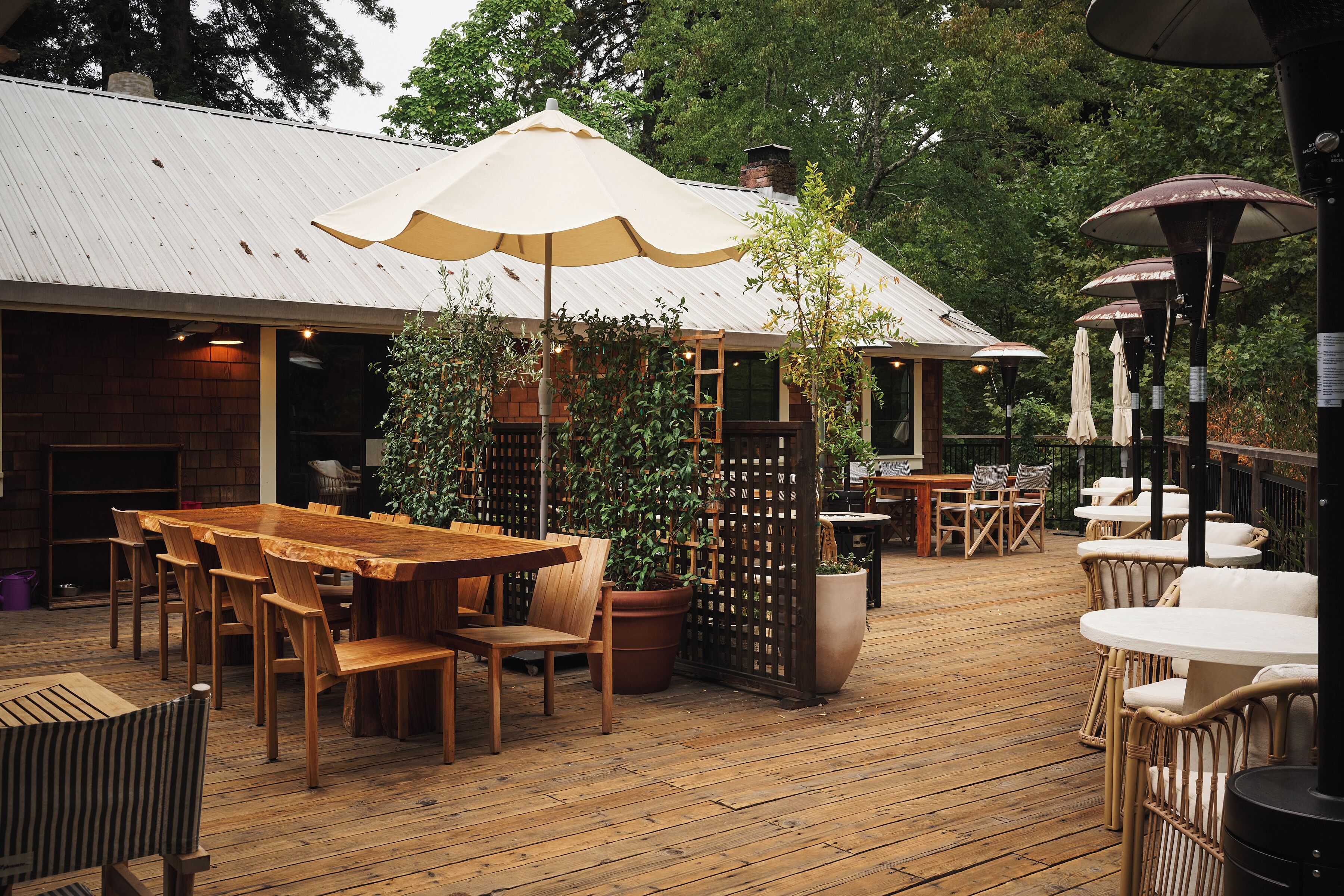 An outdoor wooden deck with tables and chairs, surrounded by potted plants and umbrellas.