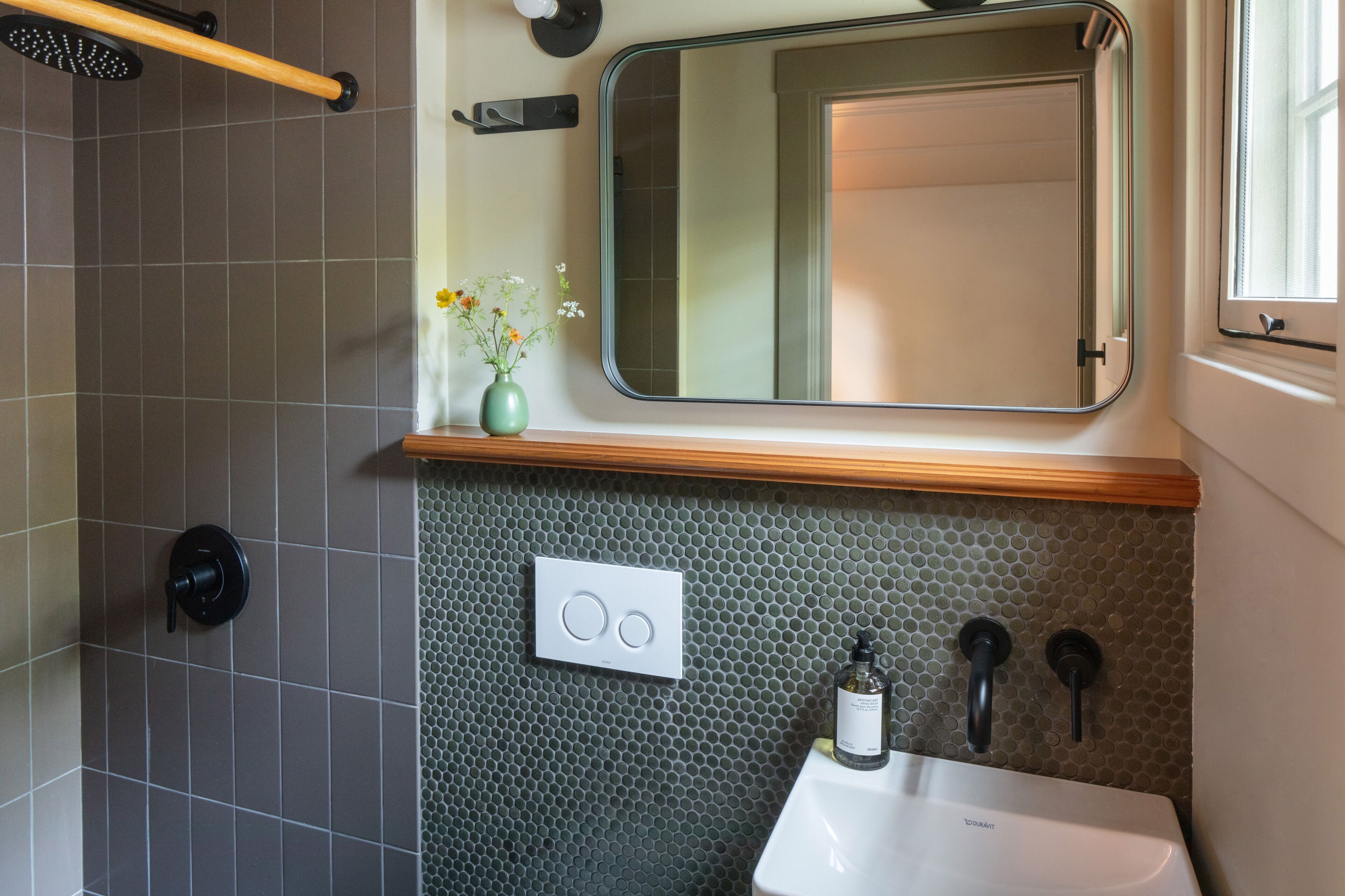 A bathroom with a shower, a sink, a mirror, and a small vase with flowers.