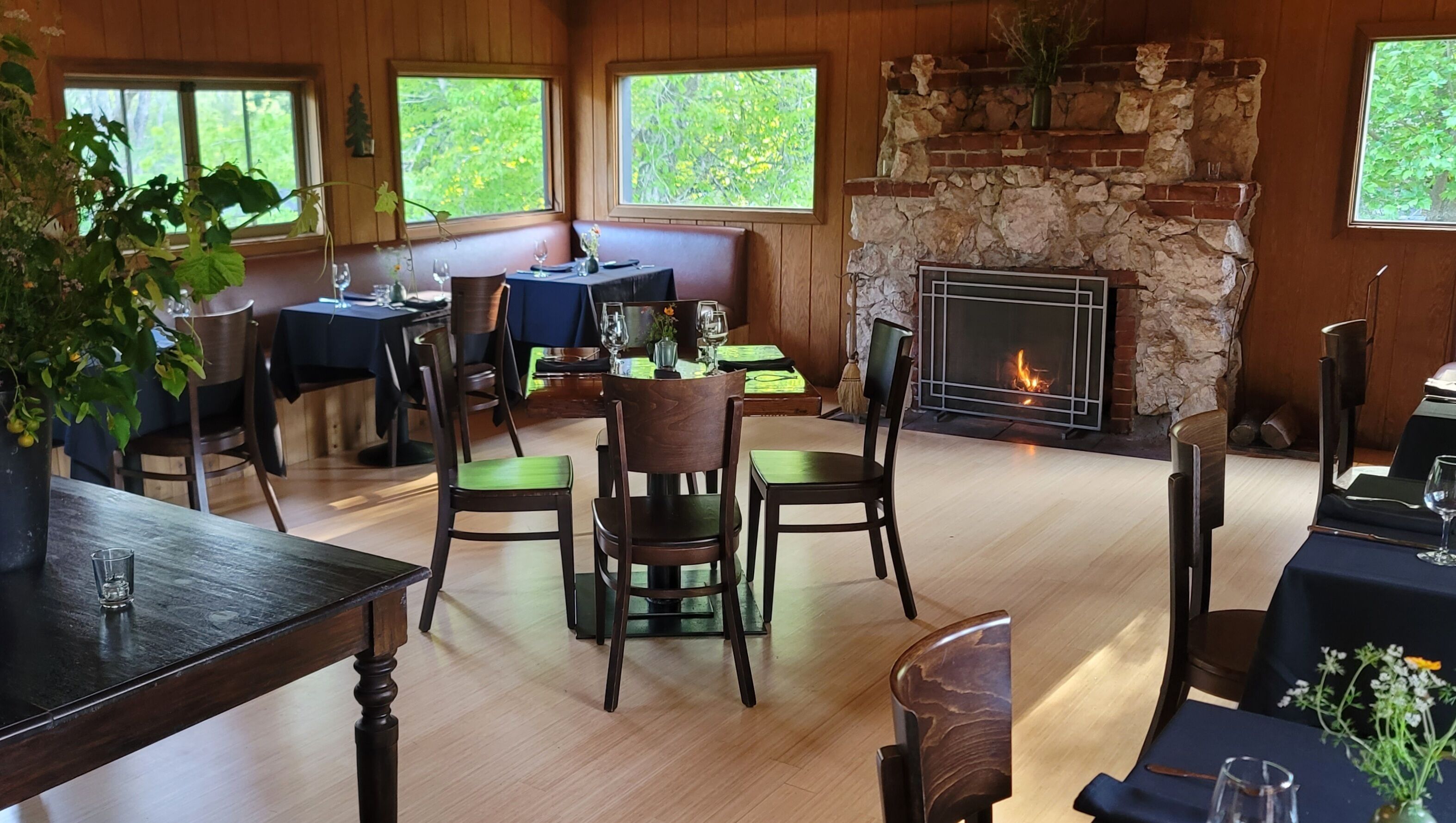 A cozy dining area with wooden walls, a stone fireplace, and large windows offering a view of greenery.