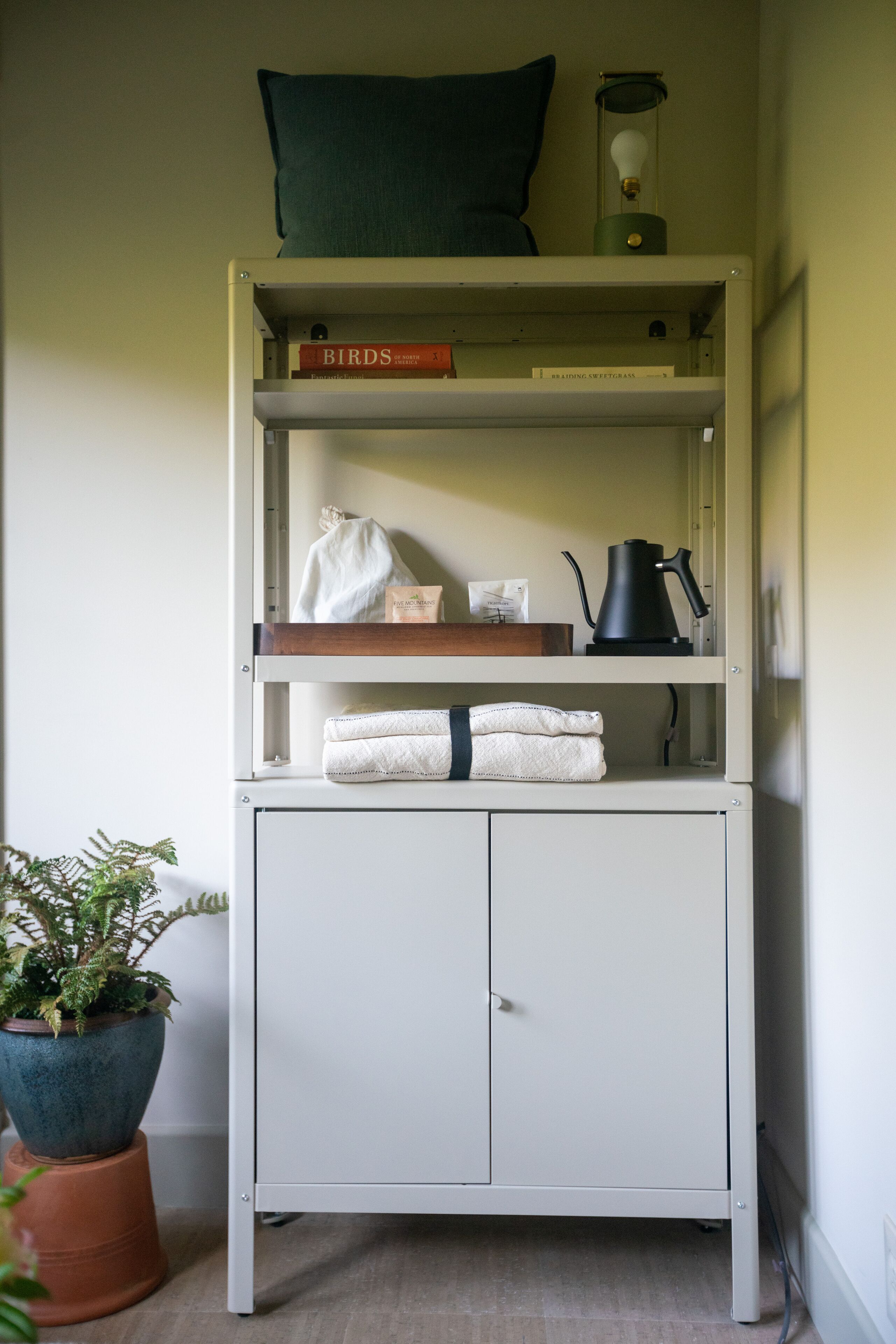 A white metal shelf unit with a green pillow, a light bulb, and books.