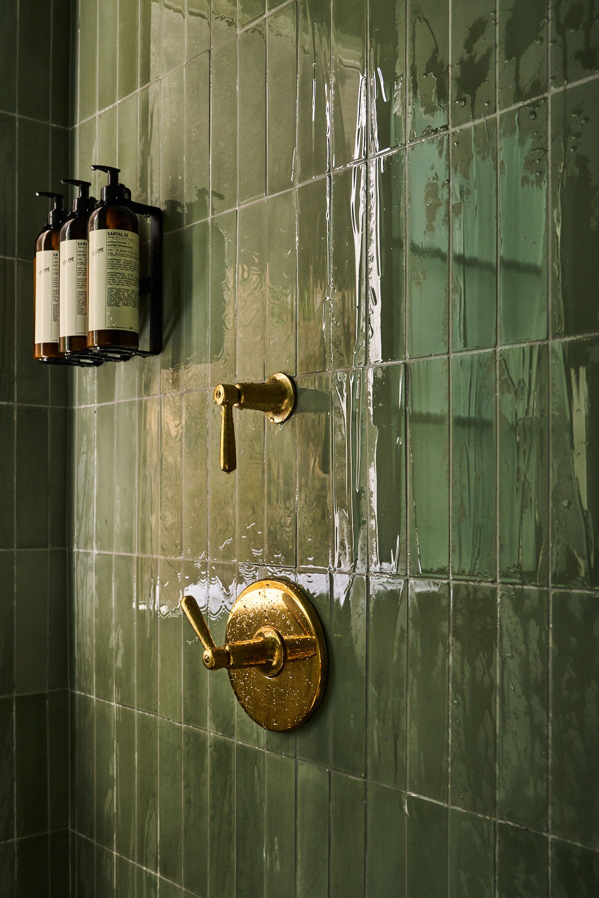A bathroom with green tiles, a gold faucet, and three bottles of Santal 33 soap.