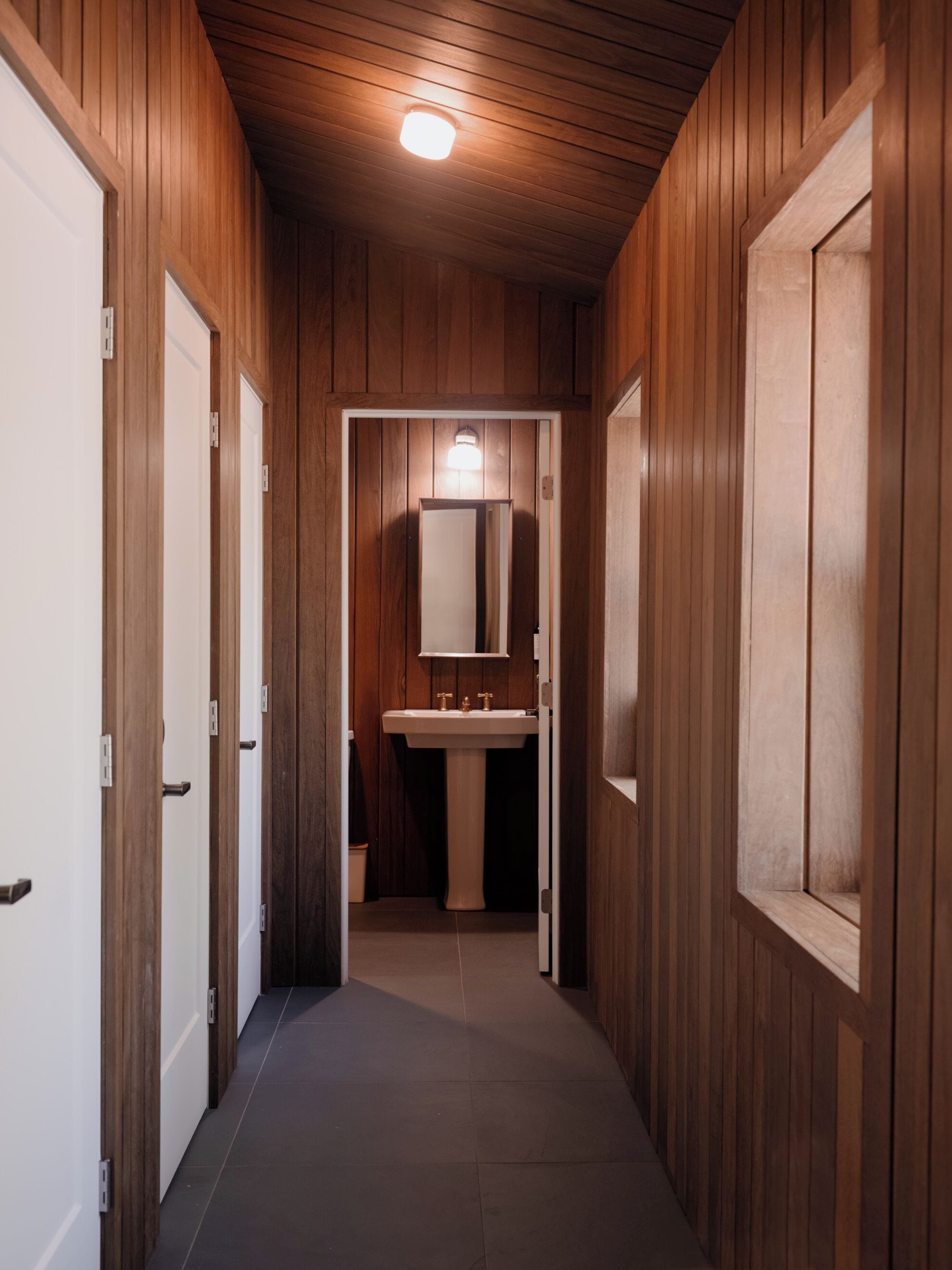 A hallway with wooden walls and doors, leading to a bathroom with a sink and mirror.