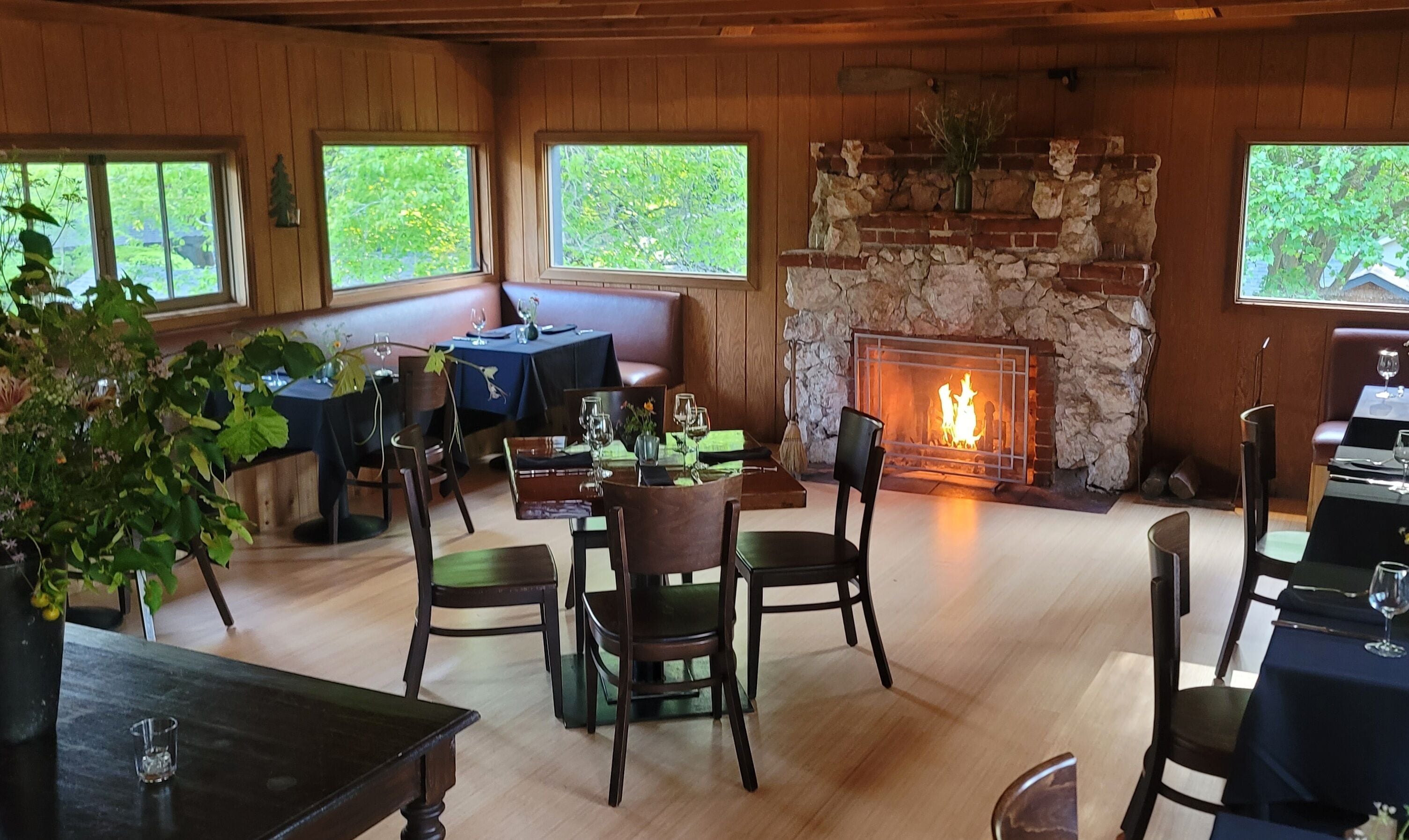A cozy dining area with wooden walls, a stone fireplace, and natural light from windows.
