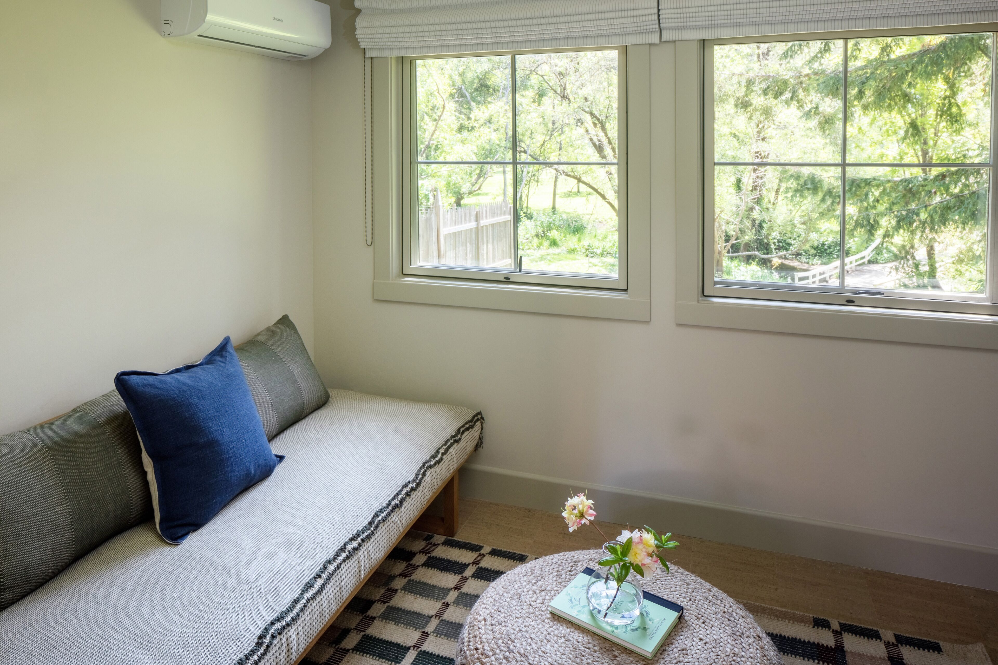 Una habitación con sofá, una mesita redonda con un libro y flores, y dos ventanas con vistas a zonas verdes.