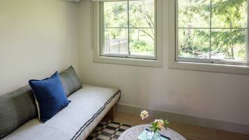 Una habitación con sofá, una mesita redonda con un libro y flores, y dos ventanas con vistas a zonas verdes.