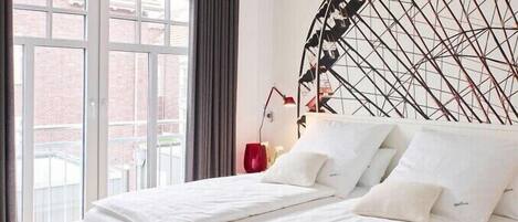 Un dormitorio moderno con una cama grande, una obra de arte geométrica en la pared y vista a un edificio de ladrillo por la ventana.