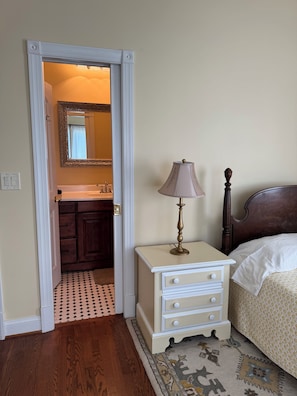 A bedroom with a bed, a nightstand, a lamp, a dresser, a mirror, and a bathroom visible through an open door.