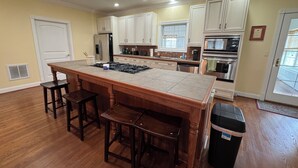 Una cocina con una isla central, taburetes de bar, refrigerador, estufa, armarios y un cubo de basura.